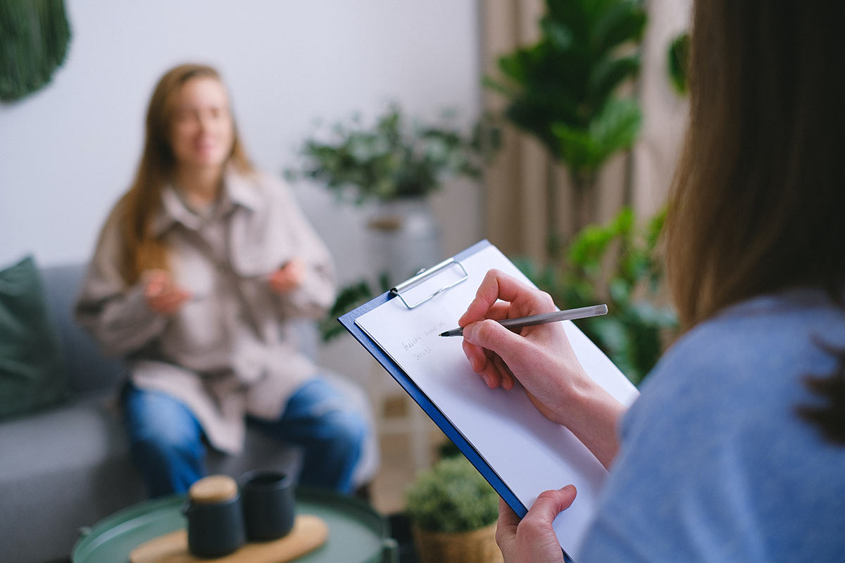 A therapist takes notes while actively listening to a smiling client, a process essential for understanding her story and creating an effective treatment path.
