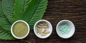 Kratom leaves with powder, capsules, and tablets displayed on a wooden surface, showing the different forms in which kratom is sold.